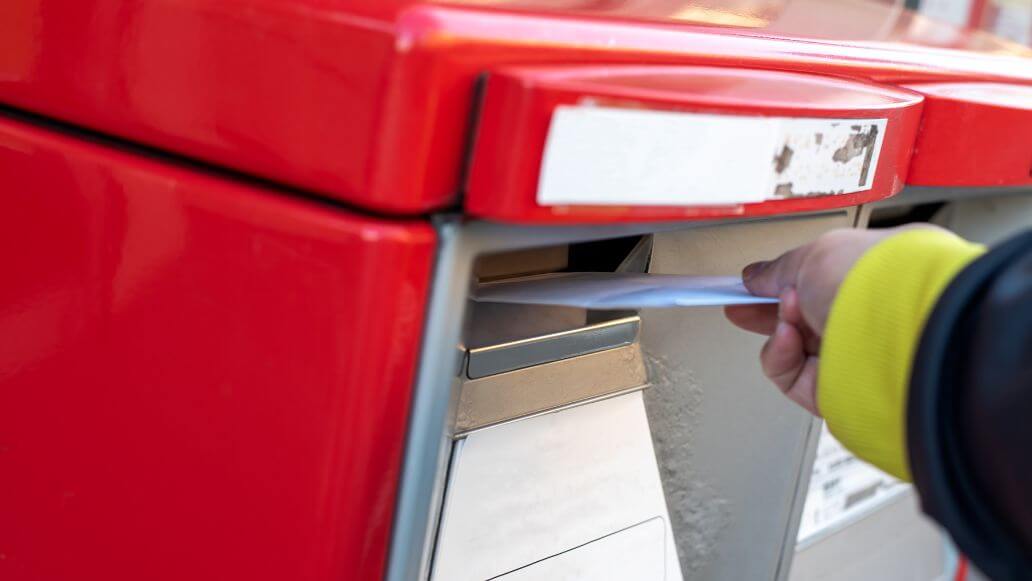 赤いポストの郵便箱に封筒を落とす手
